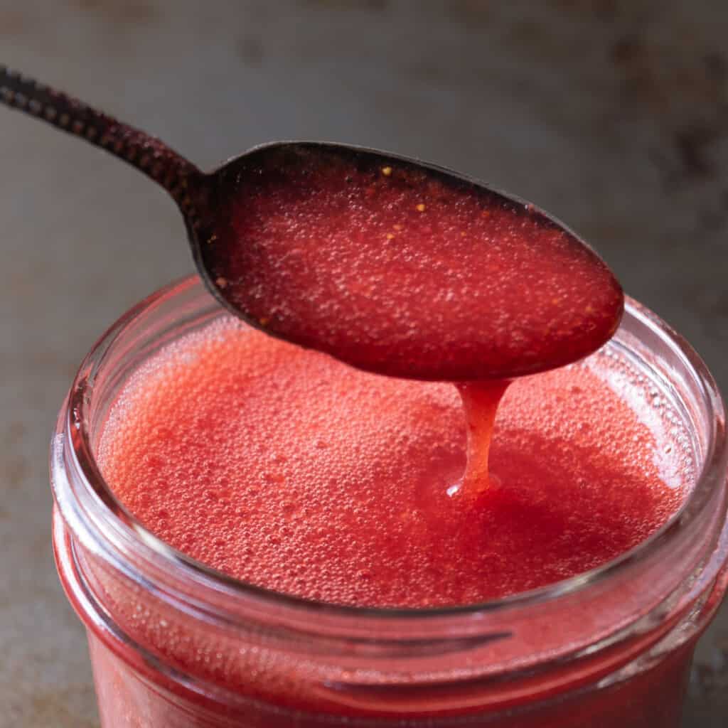 A spoonful of strawberry puree dripping into a jar.