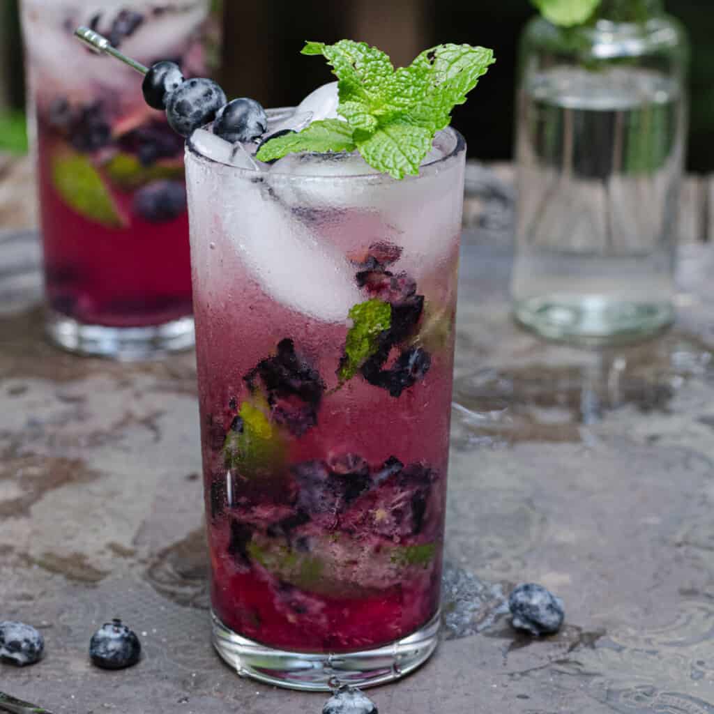 A tall blueberry mojito mocktail garnished with fresh blueberries on a serving tray outside.