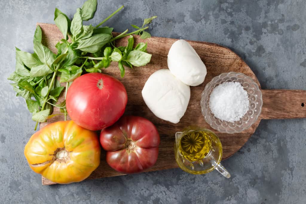 Ingredient for making caprese: heirloom tomatoes, fresh basil, mozzarella, extra virgin olive oil, and salt flakes.