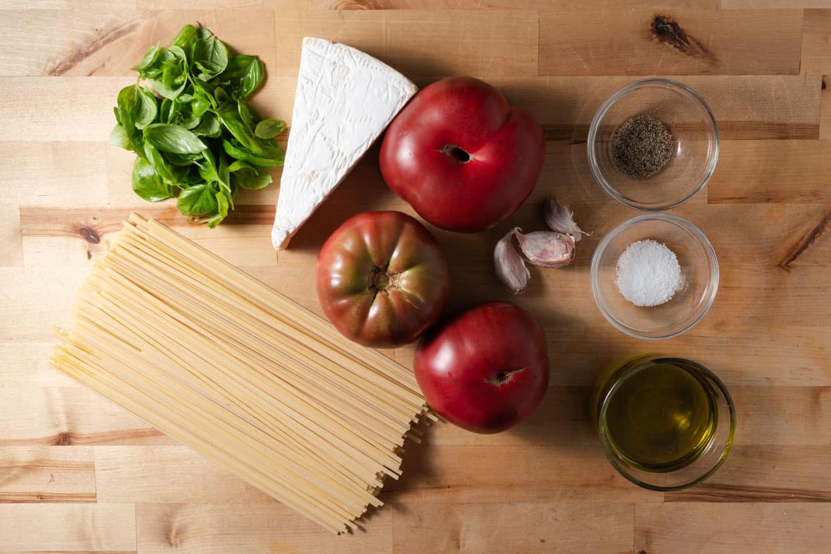 Dried linguine, whole heirloom tomatoes, a wedge of brie cheese, fresh basil, garlic cloves, extra virgin olive oil, black pepper, and kosher salt on a cutting board.