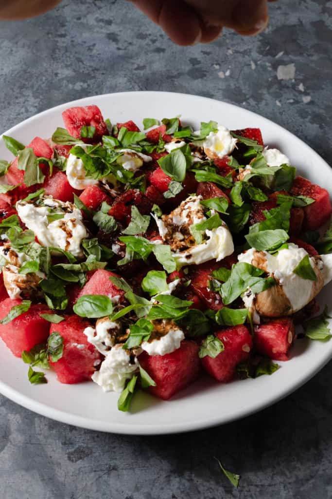 Adding salt flakes to a summer burrata salad with fresh melon, herbs, and balsamic.