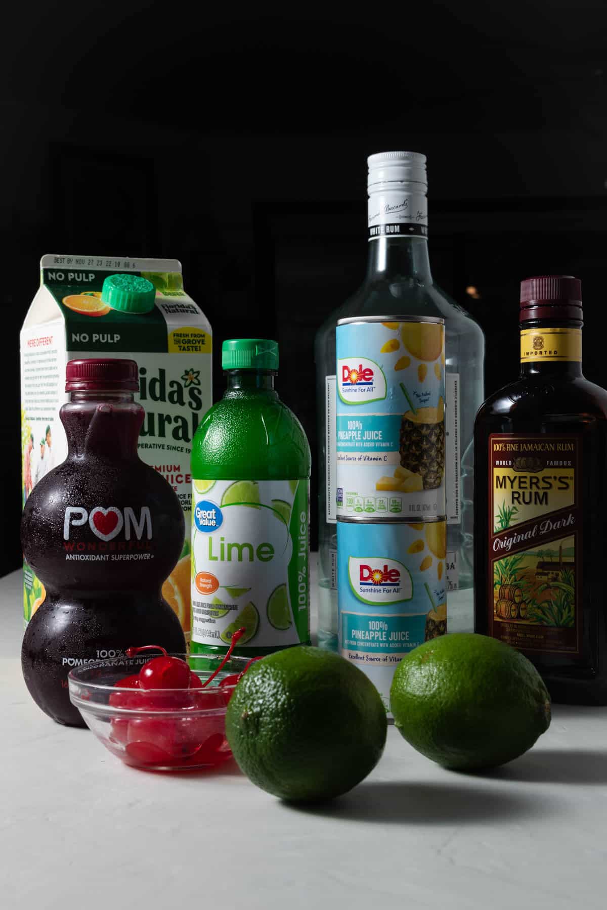 Ingredients for making rum punch with limes, lime juice, maraschino cherries, orange juice, pineapple juice, pomegranate juice, lime juice, Bacardi rum, and Myers rum.