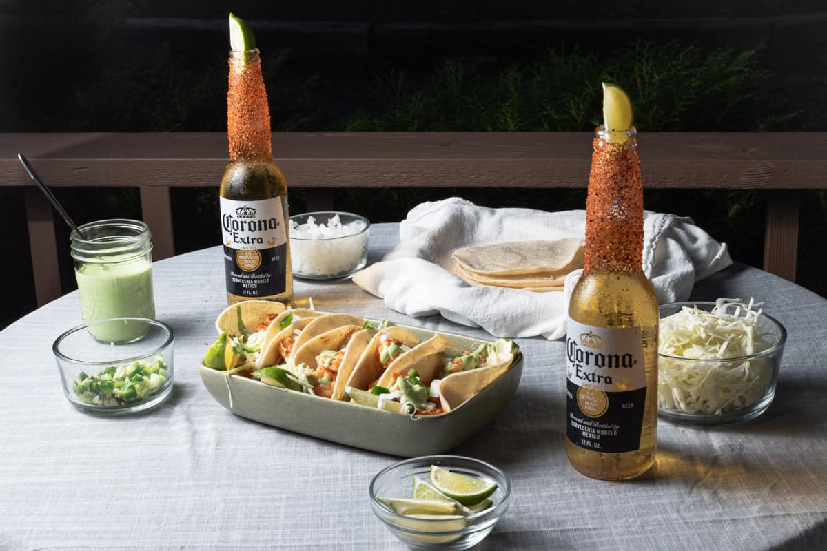 Chili lime shrimp tacos with cilantro lime crema, onions, and jalapenos on corn tortillas on an outdoor table with toppings and dressed beer.