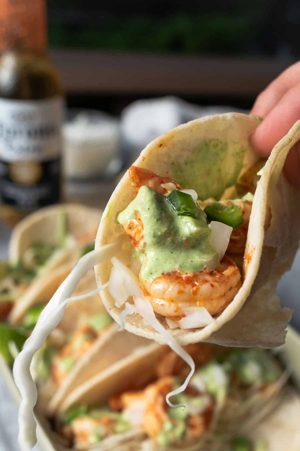 A hand holding a shrimp taco with tacos and beer in the background.