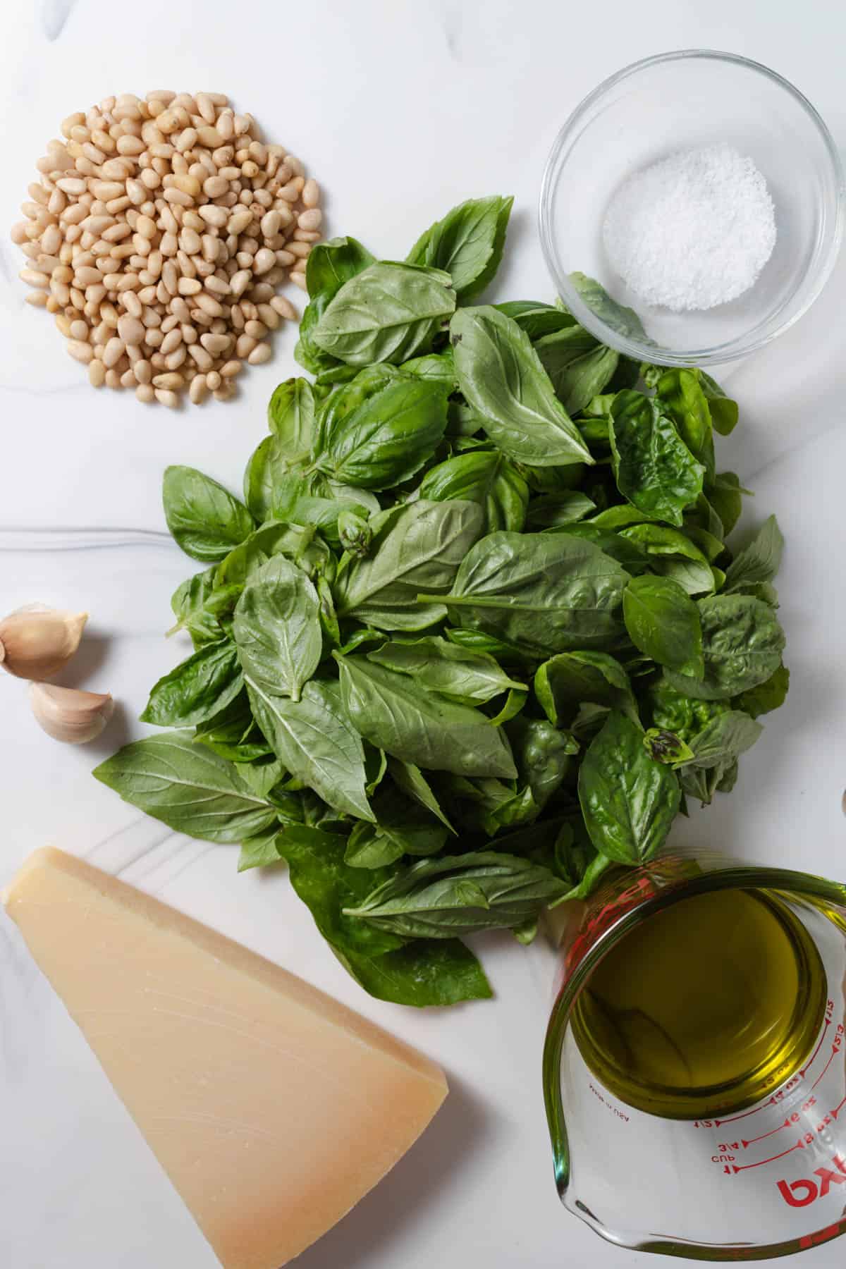 Fresh basil leaves, garlic, parmesan cheese, extra virgin olive oil, pine nuts, and salt on a cutting board.