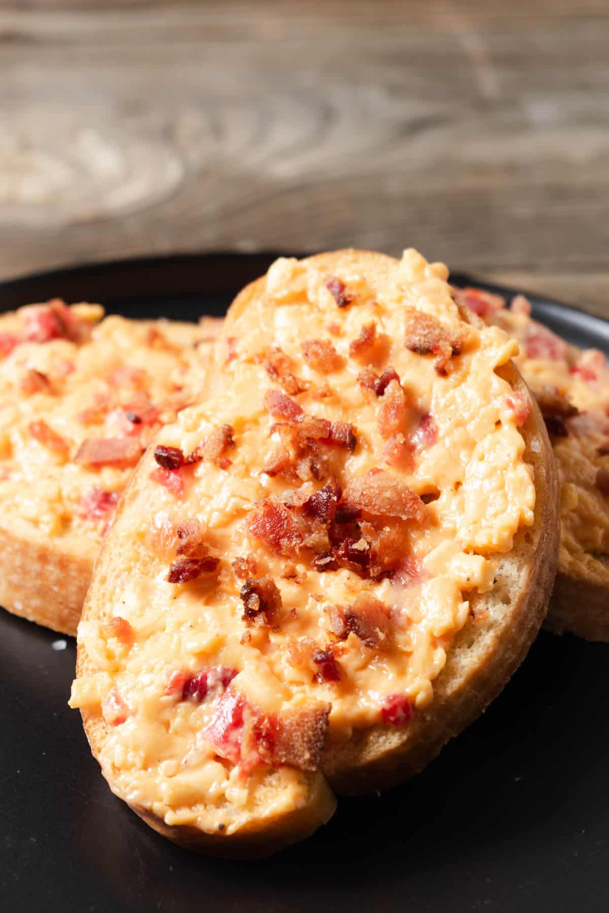 Pimento cheese and bacon crostini served on a black cocktail plate.
