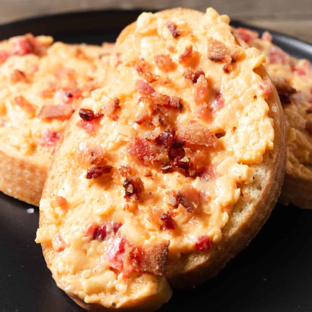 Pimento cheese and bacon crostini served on a black cocktail plate.