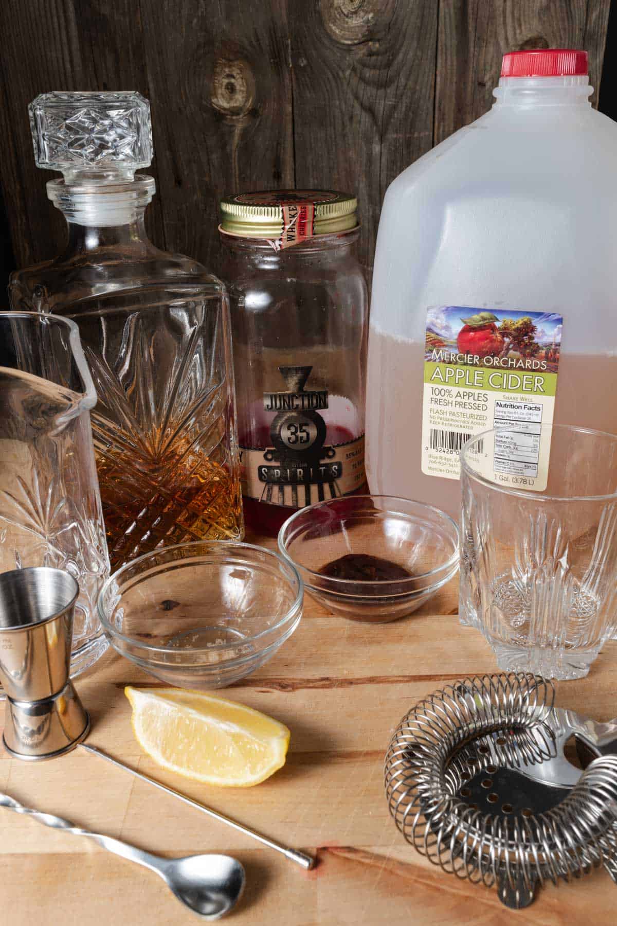 Bourbon, cherries, apple cider, lemon, apple butter, brown sugar, and cocktail mixing accessories laid out for drink mixing.