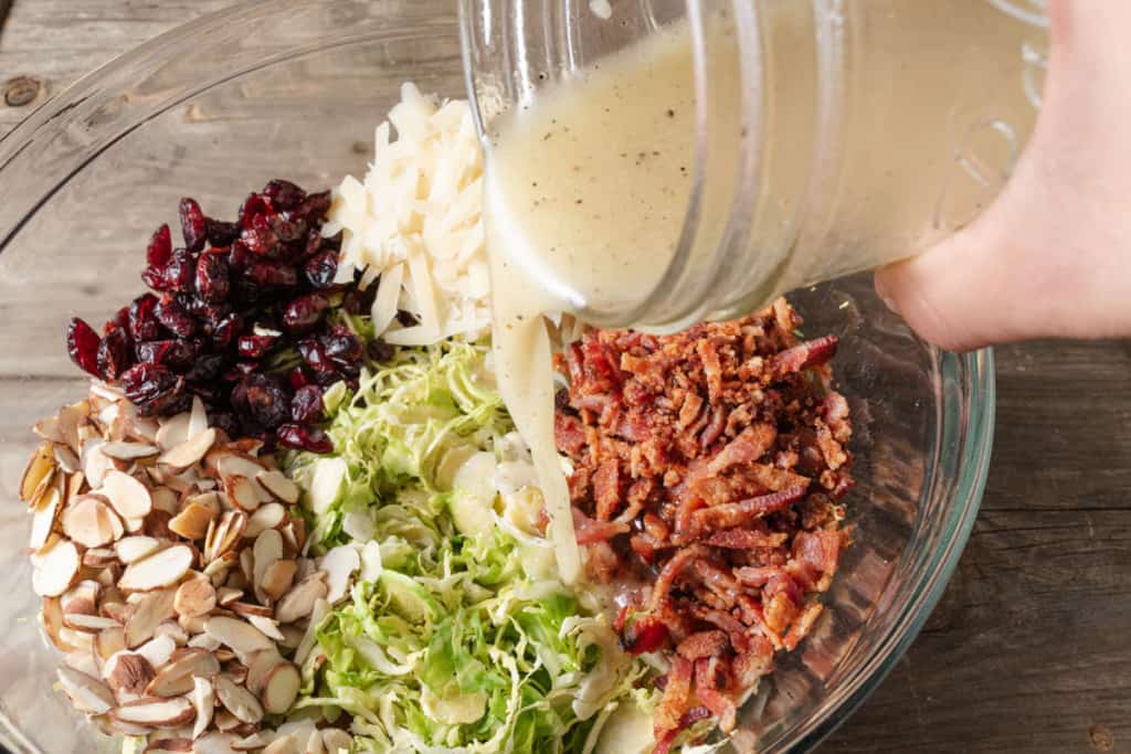 Pouring a lemon vinaigrette dressing into a bowl of shaved brussels sprouts, almonds, cranberries, bacon, and parmesan.