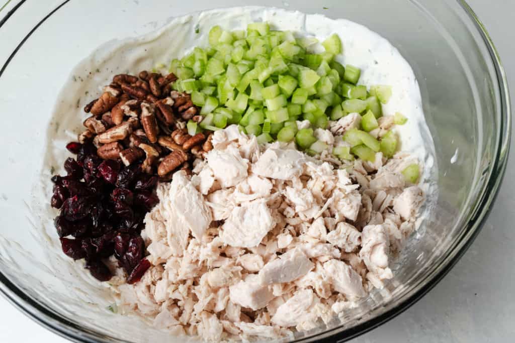 A glass bowl with chopped chicken breast, dried cranberries, celery, pecan pieces, and tangy herb dressing.