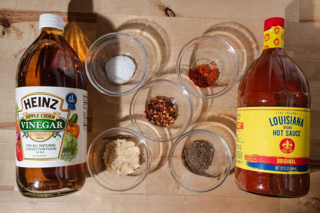 Cider vinegar, hot sauce, brown sugar, pepper flakes, salt, pepper, and cayenne on a butcher block.