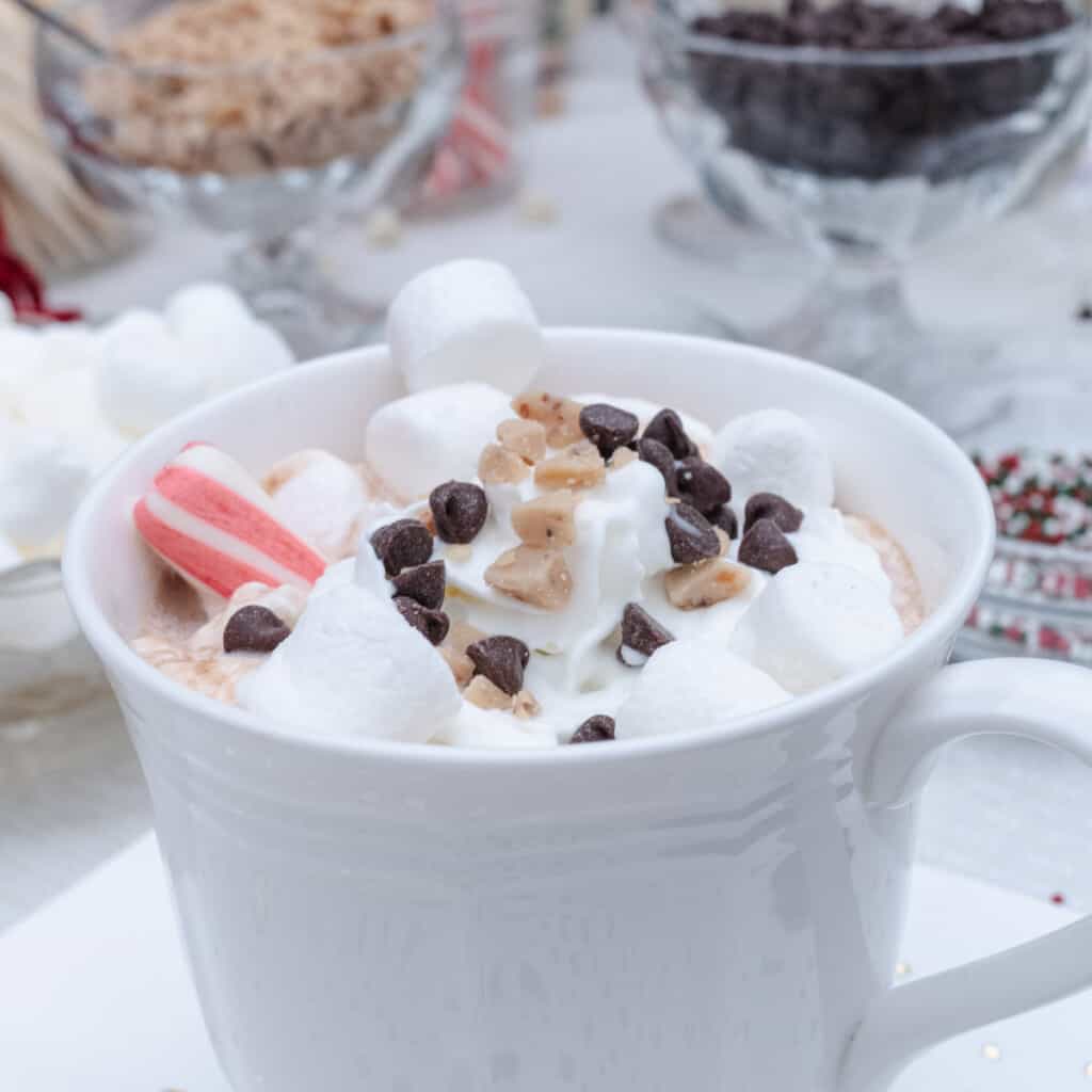 A mug of hot chocolate at a hot chocolate bar with marshmallows, toffee bits, chocolate chips, whipped cream, and a peppermint stick.