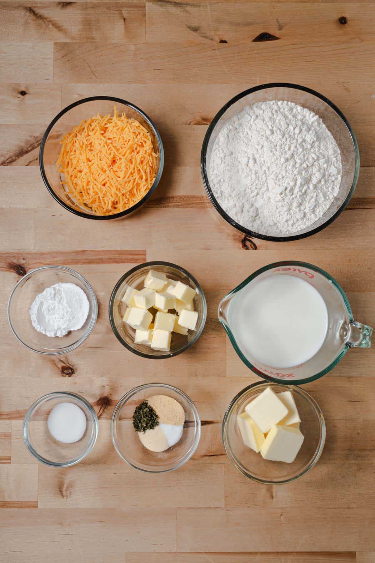 All purpose flour, baking powder, salt, butter, shredded cheddar cheese, milk, garlic, and herbs measured out to make cheddar bay drop biscuits.