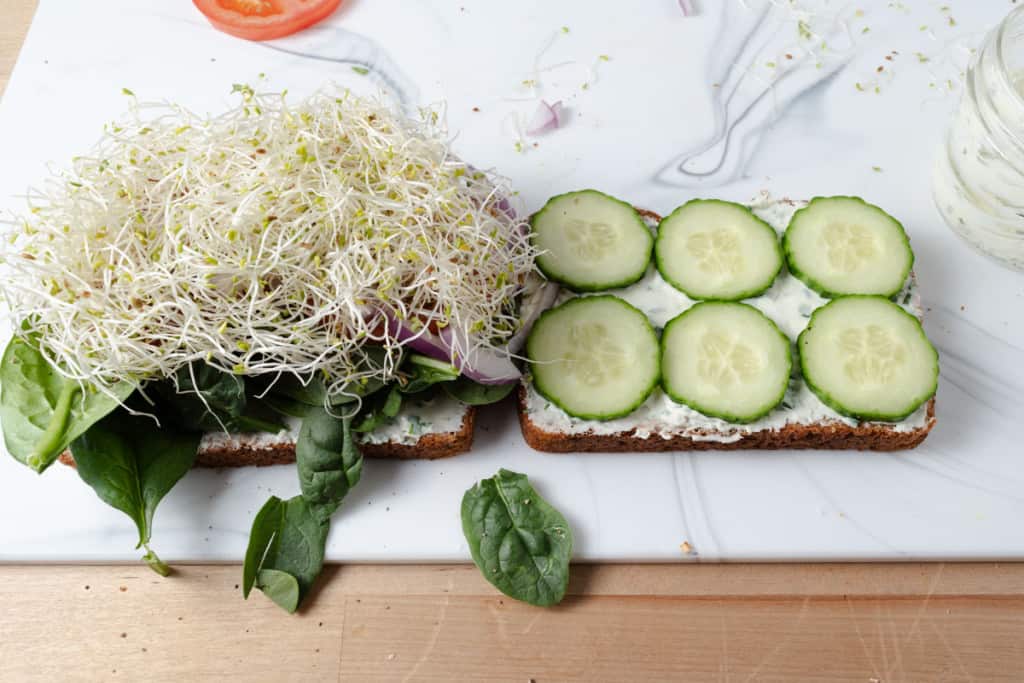 Adding alfalfa sprouts and cucumber slices to a cream cheese and vegetable sandwich.