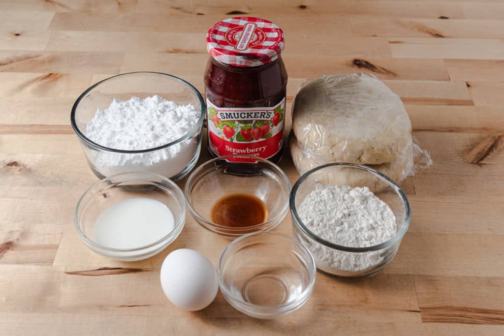 Pie dough, strawberry jam, powdered sugar, milk, vanilla extract, flour, and an egg on a countertop.