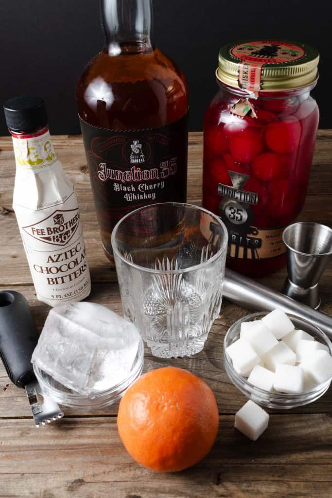 Black cherry whiskey, chocolate bitters, sugar cubes, bourbon cherries, an orange, a large cube of ice, and an orange zester.