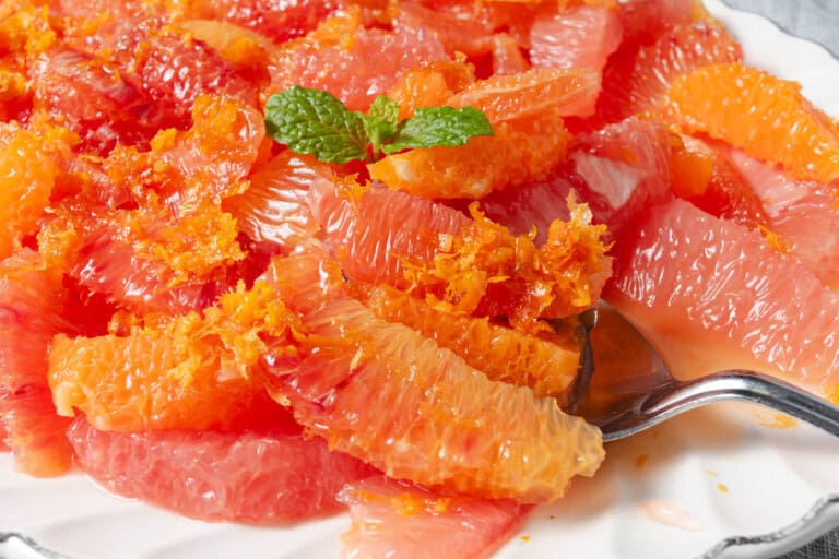 A close-up of a grapefruit salad with vibrant pink and orange segments, garnished with a sprig of fresh mint. The dish is served on a white plate, with a silver serving spoon on the side.