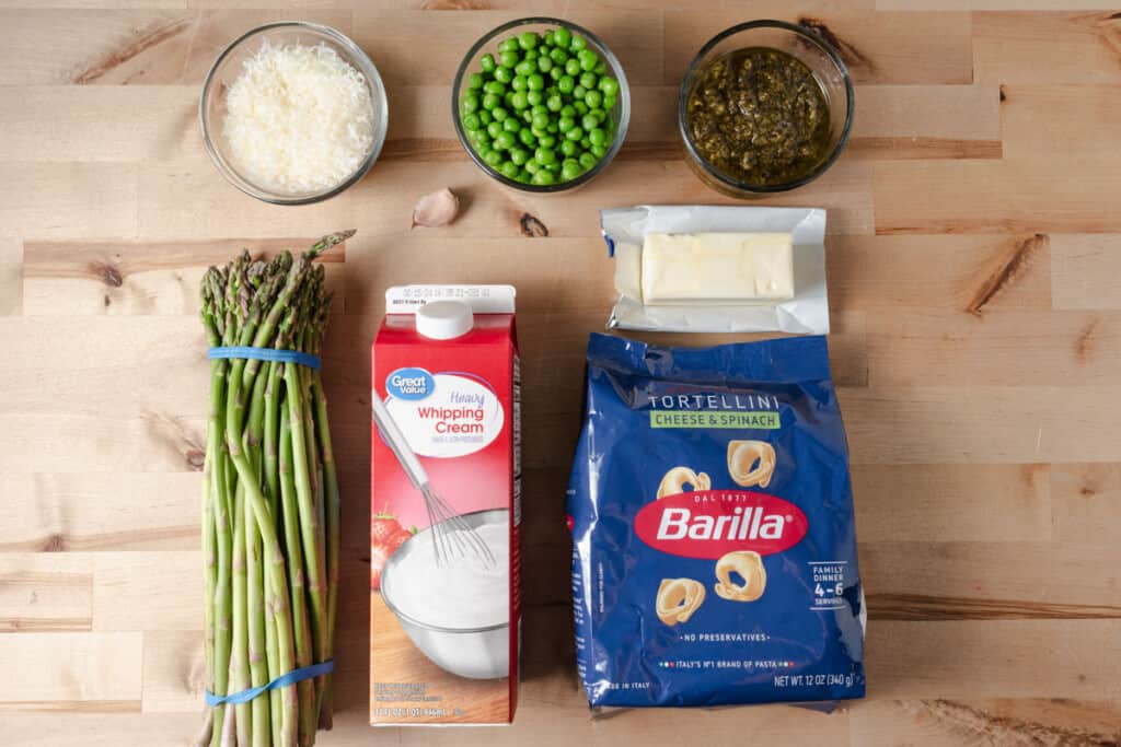 Dried tortellini, heavy cream, butter, garlic, pesto, parmesan cheese, peas, and asparagus on a countertop.