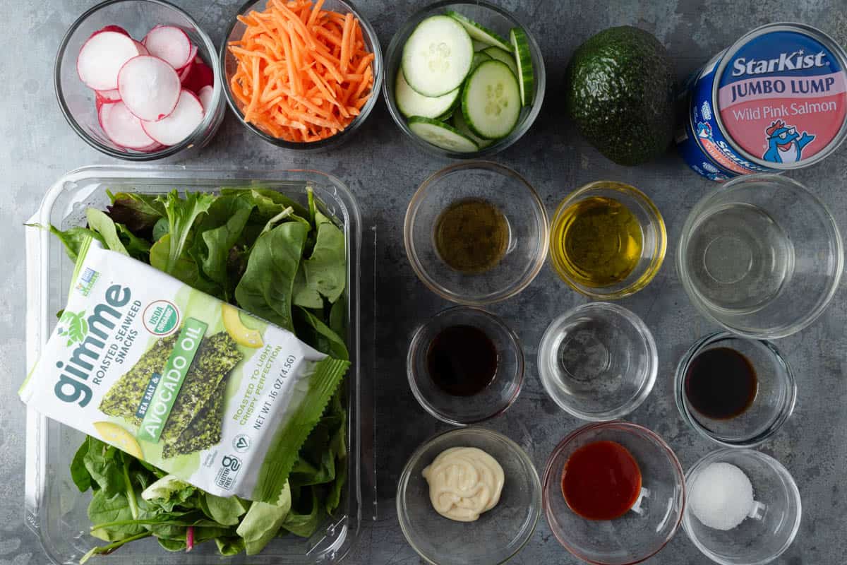 Mixed greens, seaweed snacks, radishes, shredded carrots, cucumbers, avocado, tinned salmon, sriracha, kewpie mayo, sugar, sesame oil, olive oil, and soy sauce measured out to make an easy salad.