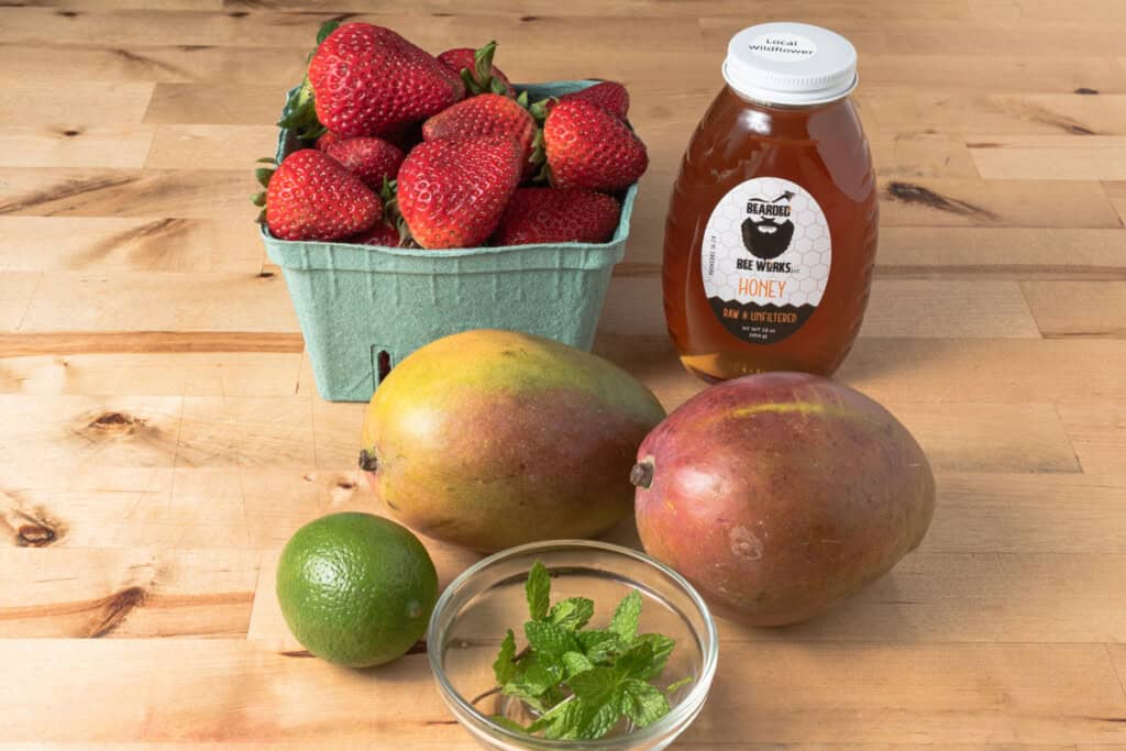 A table with a bottle of honey, strawberries, mangoes, a lime, and a bowl of mint.