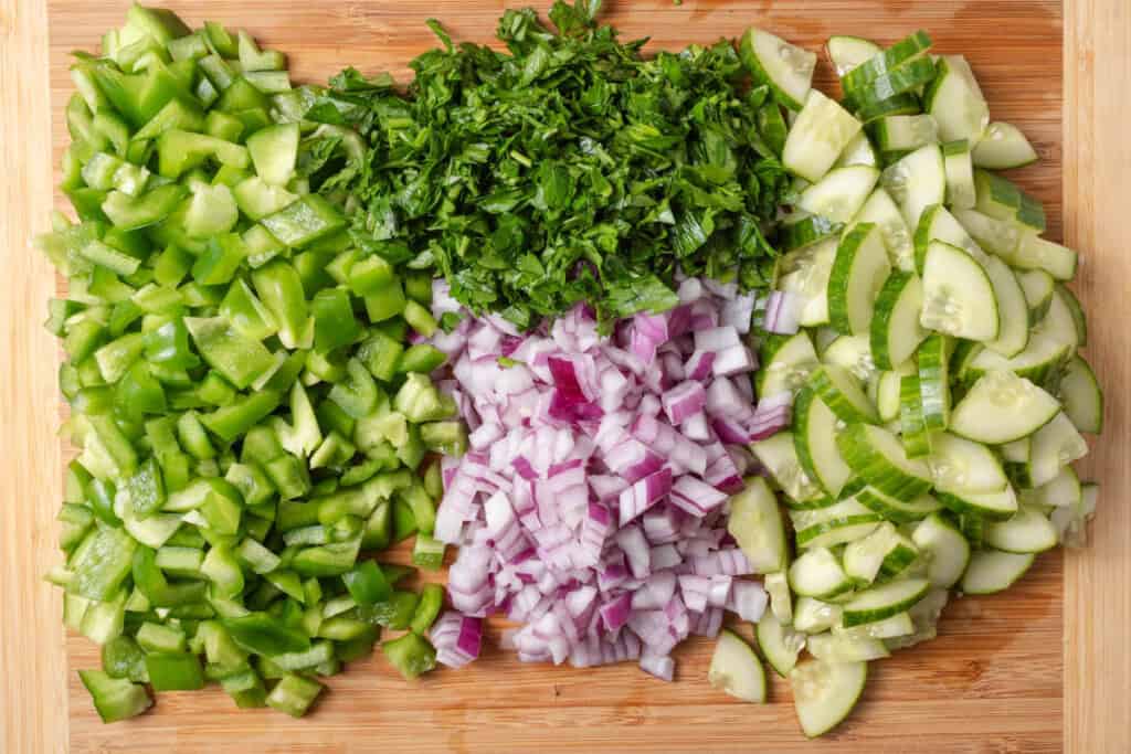 Chopped bell peppers, cucumbers, red onions, and parsley on a wooden cutting board.