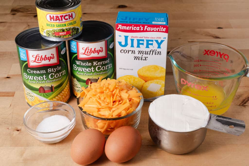 Jiffy corn muffin mix, canned corn, eggs, cheese, sour cream, sugar, green chiles, and melted butter on a countertop.