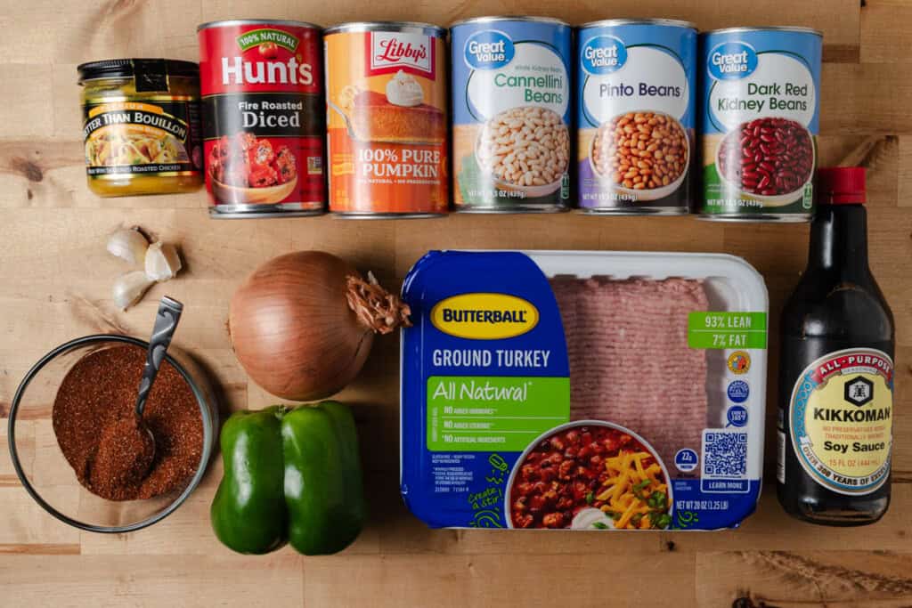 Ground turkey, homemade chili seasoning, soy sauce, chicken bullion, bell pepper, yellow onion, garlic cloves, fire roasted tomatoes, pumpkin puree, cannellini, pinto beans, and dark red kidney beans on a countertop.