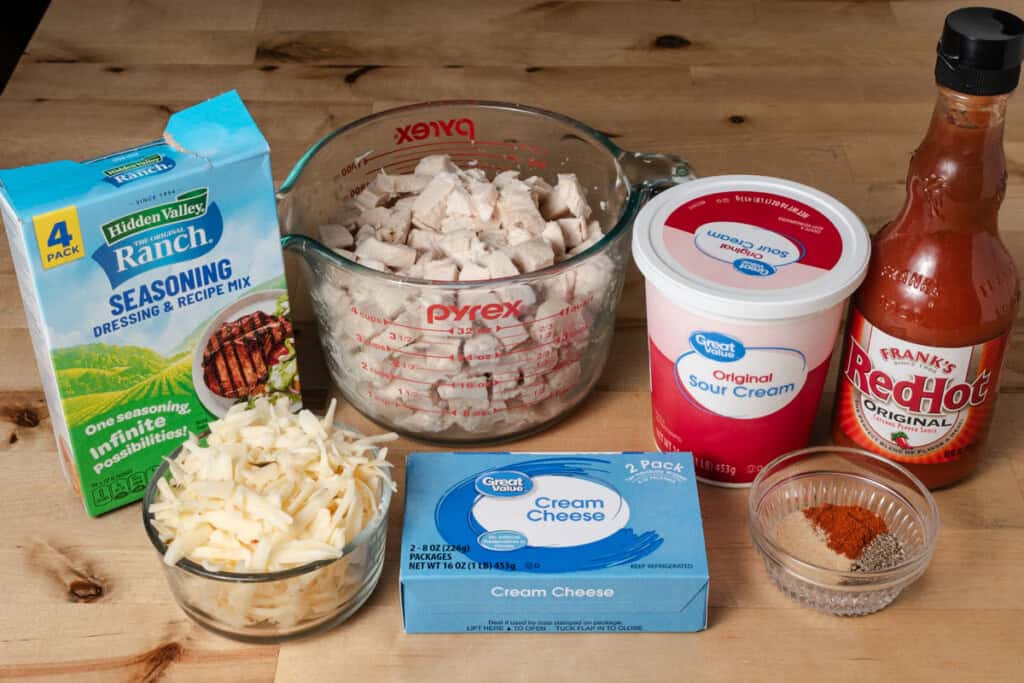 Cooked chicken breast, Frank's RedHot sauce, ranch seasoning, cream cheese, sour cream, pepper jack cheese, garlic powder, cayenne, and black pepper arranged on a countertop.