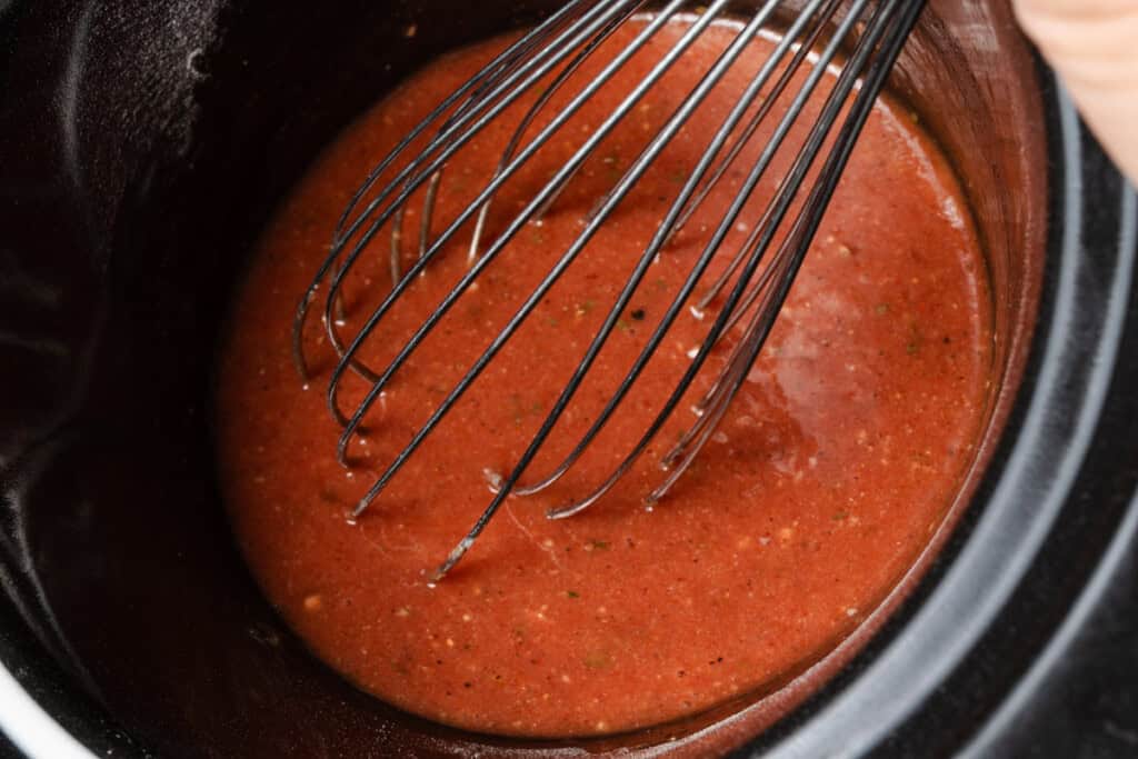 Whisking Frank's hot sauce, ranch seasoning, garlic, cayenne, and pepper in a slow cooker insert.
