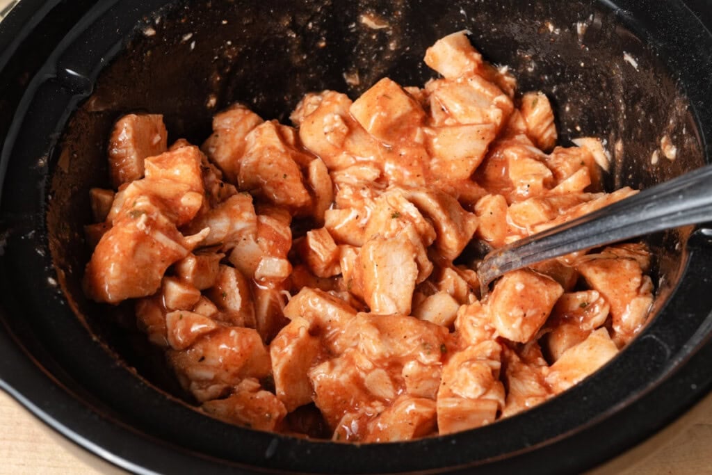 Mixing cooked chicken breast with Frank's hot sauce and ranch seasoning in a slow cooker insert.