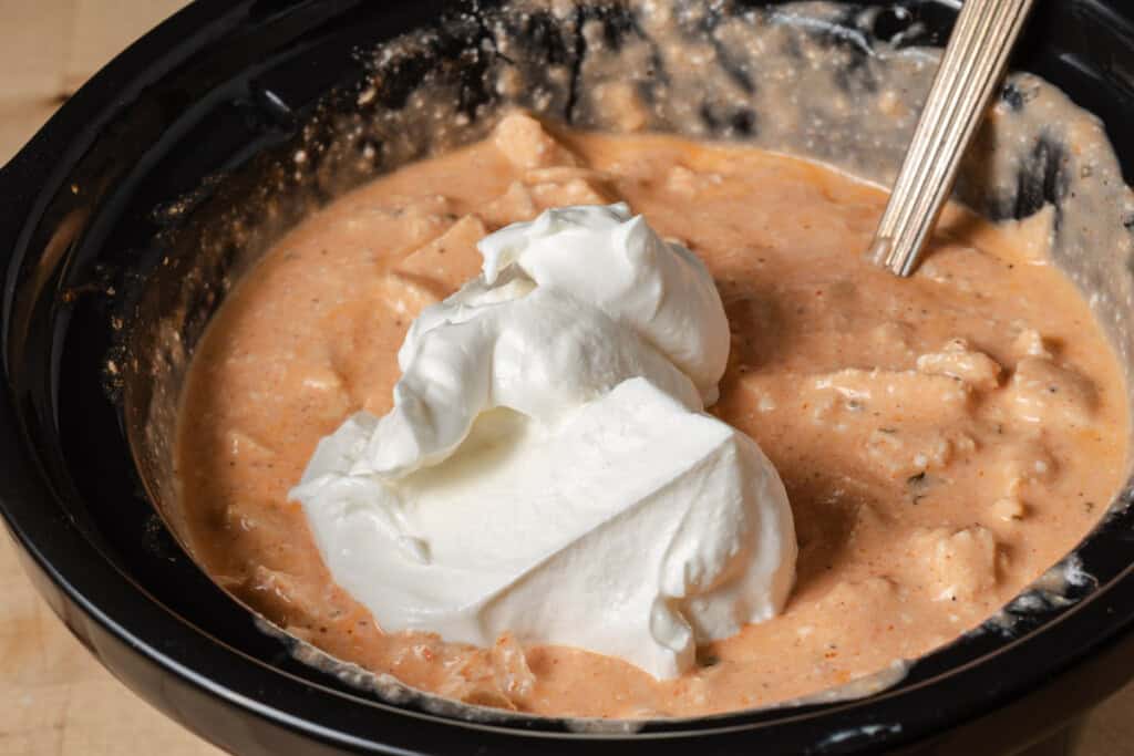 Adding the sour cream to slow cooker buffalo chicken dip.