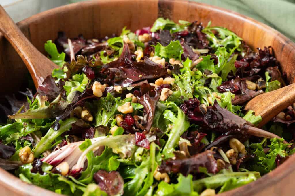 A bowl of salad greens with dried cranberries, walnuts, and balsamic vinaigrette with salad serving tongs.