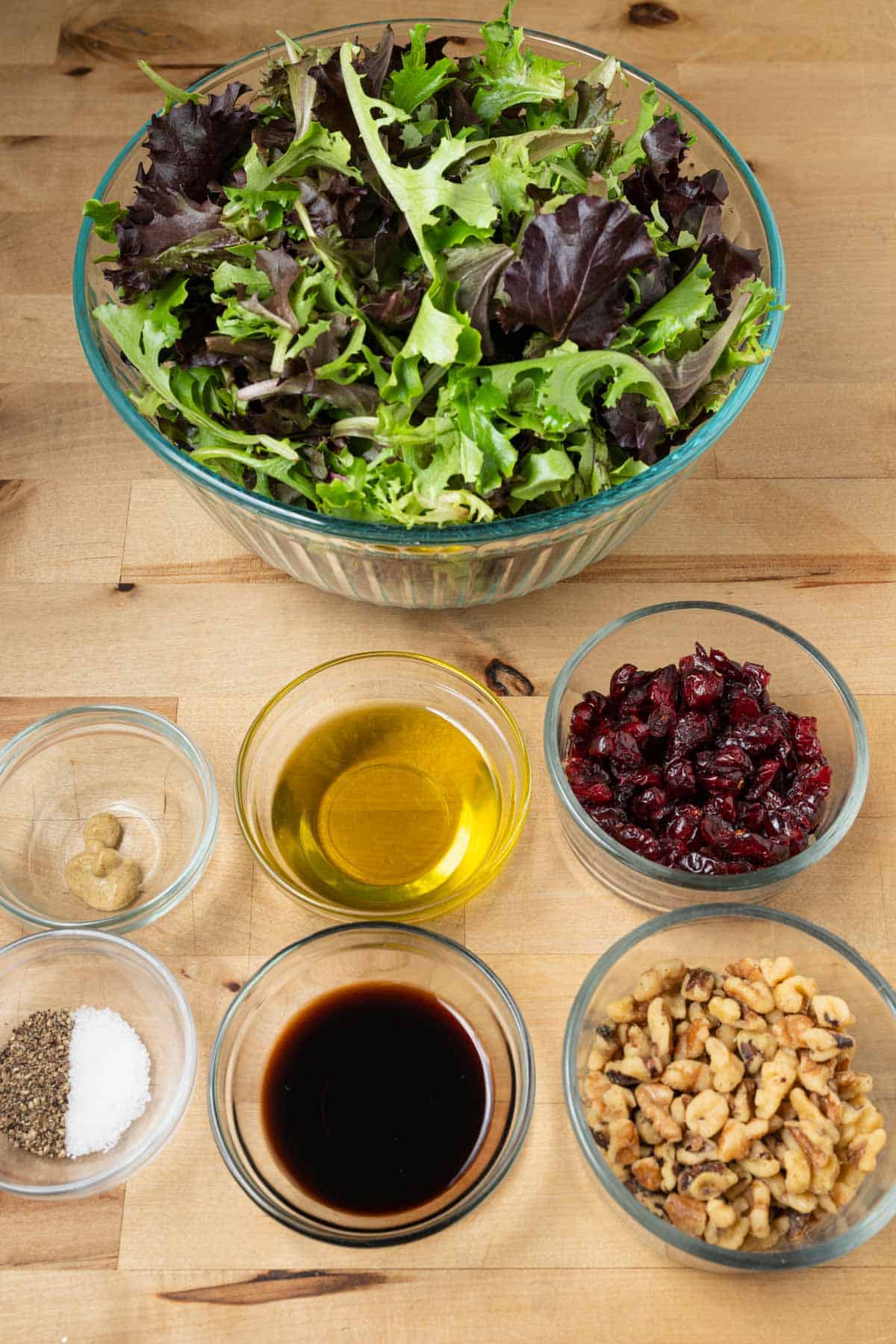 Fresh organic salad greens, dried cranberries, walnut pieces, balsamic vinegar, olive oil, Dijon mustard, salt, and pepper prepped for making a salad.