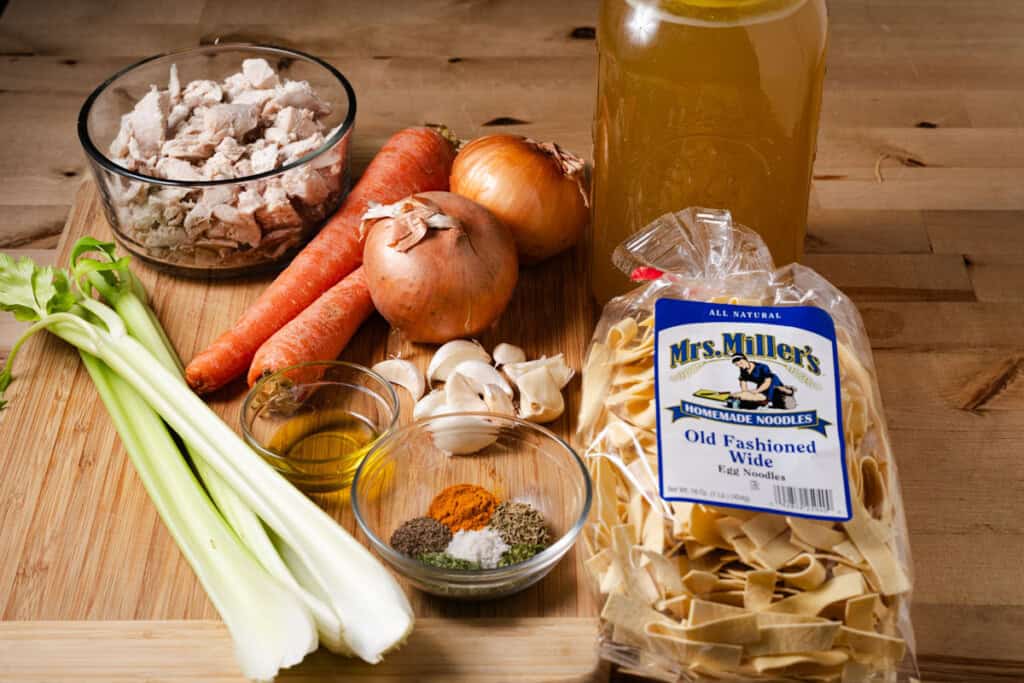 Cooked chicken, homemade stock, celery, onions, carrots, garlic, olive oil, herbs, spices, and egg noodles on a countertop.