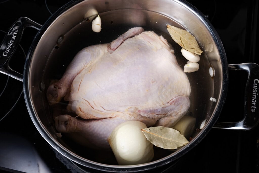 A whole chicken in a stockpot with poaching liquid and seasonings.