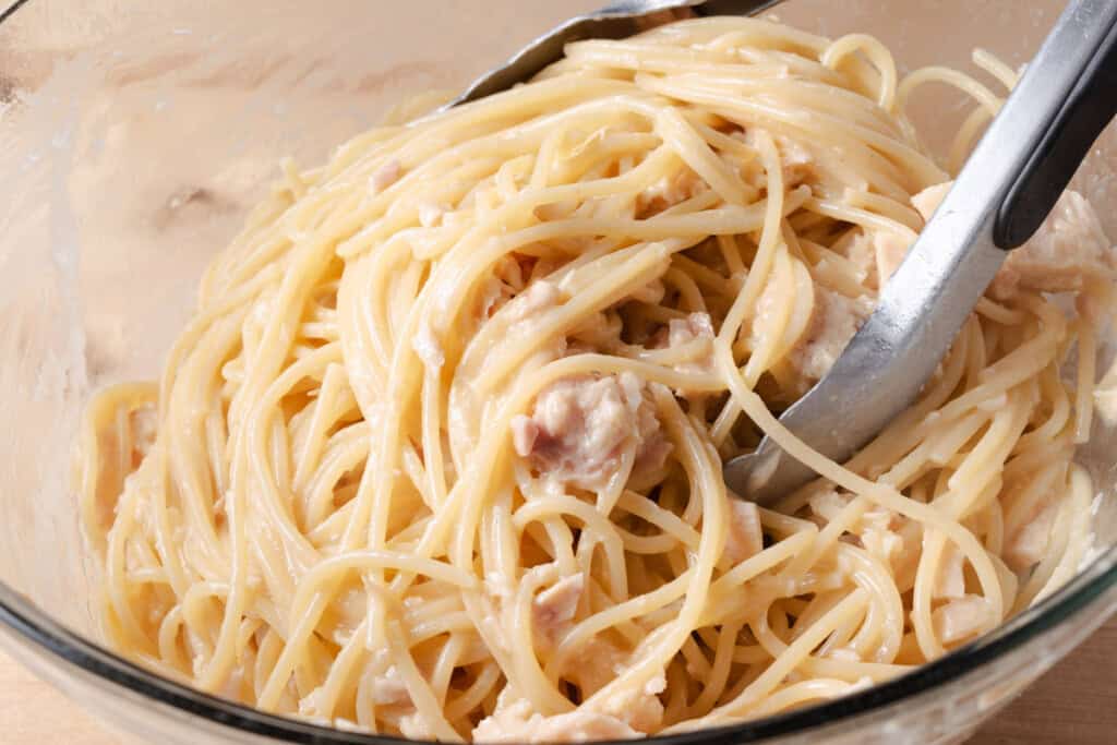 Using tongs to toss hot spaghetti noodles with tuna, olive oil, and butter.