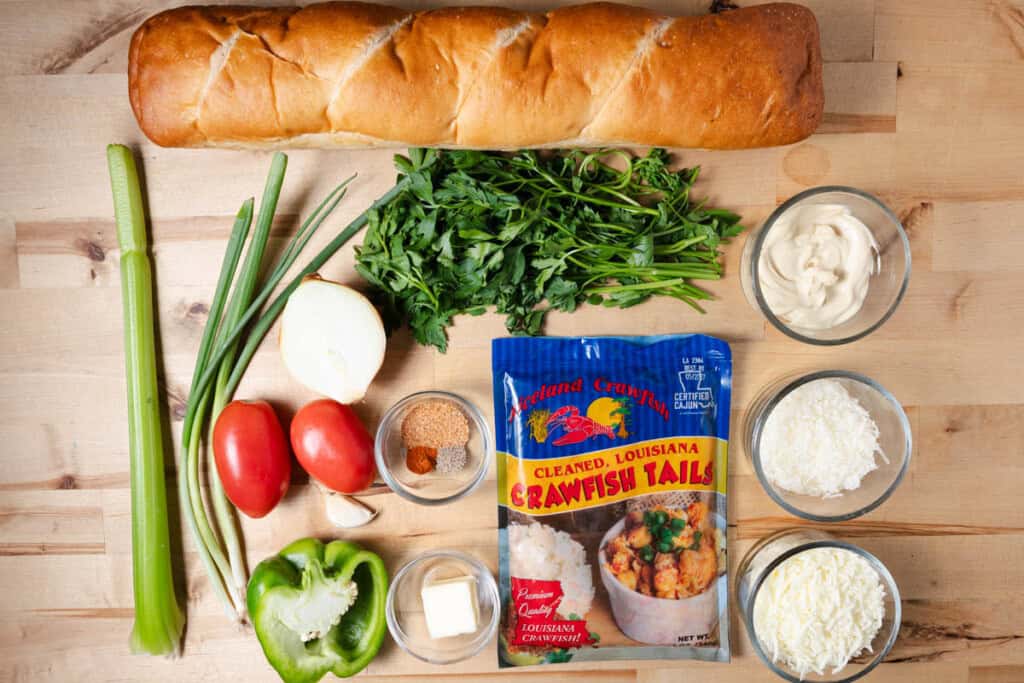 A loaf of French bread, crawfish tail meat, a celery stalk, half of a yellow onions, half of a bell pepper, roma tomatoes, a clove of garlic, fresh parsley, mayonnaise, shredded mozzarella, shredded parmesan, butter, and spices on a cutting board.