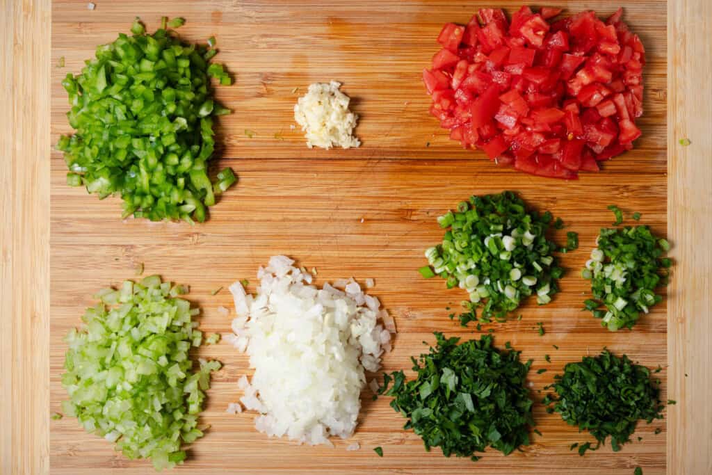 Diced celery, onions, bell peppers, tomatoes, minced garlic, chopped green onions, and chopped parsley on a cutting board.