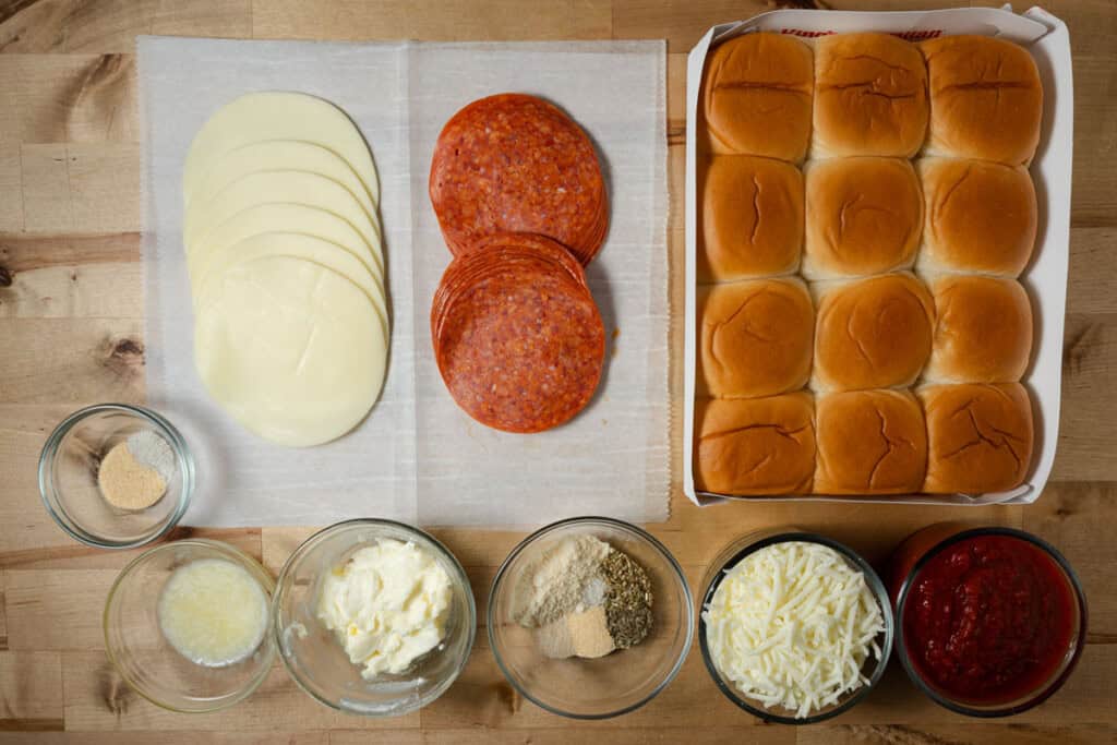A pack of King's Hawaiian rolls, slices of provolone, pepperonis, shredded mozzarella, butter, garlic, herbs, and spices, and pizza sauce on a cutting board.
