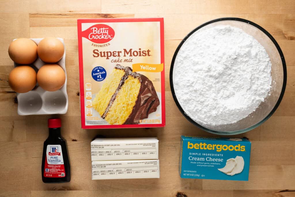 Yellow cake mix, powdered sugar, butter, cream cheese, eggs, and vanilla on a countertop.