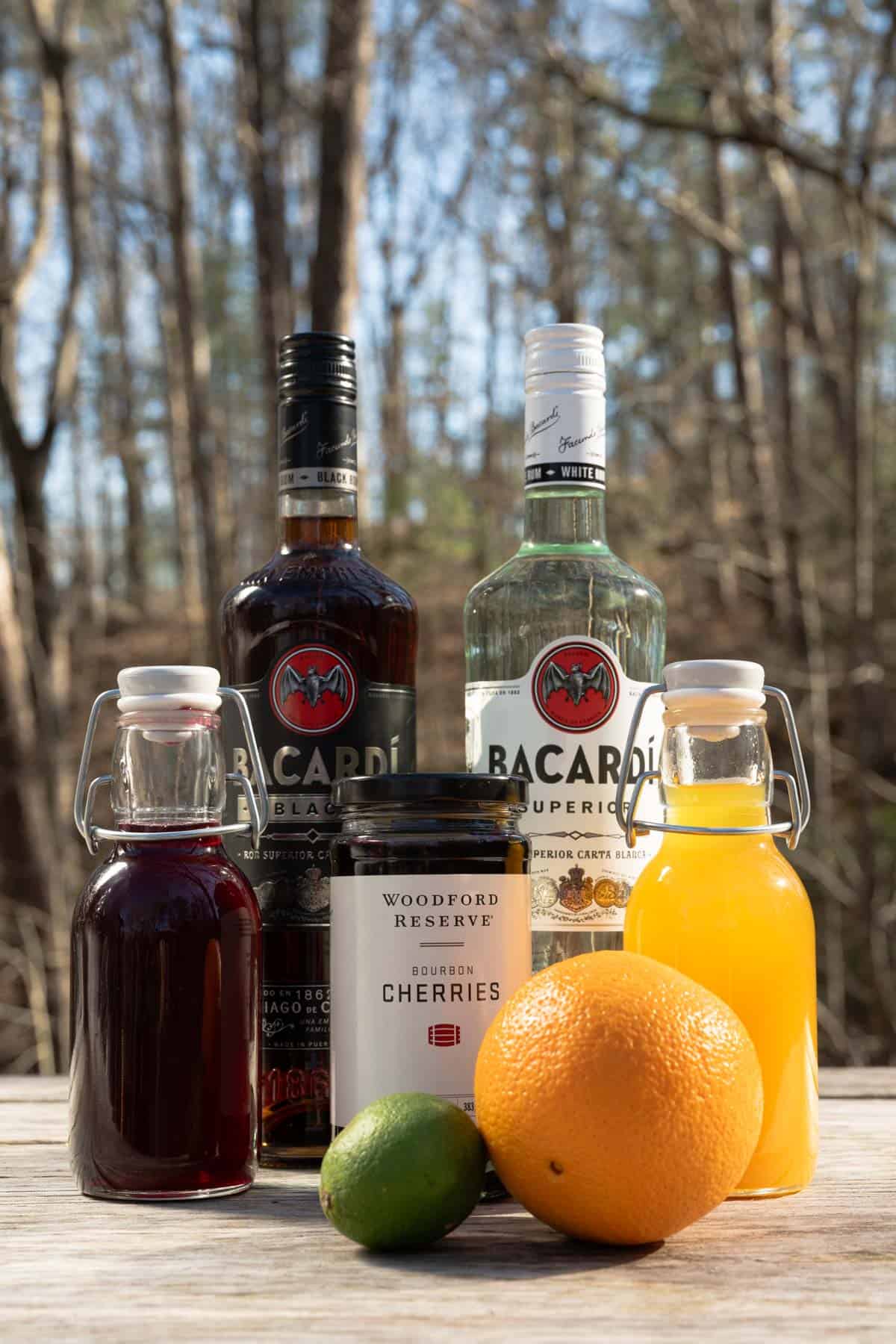 Dark rum, light rum, grenadine, passion fruit syrup, cherries, a lime, and an orange on an outdoor table.