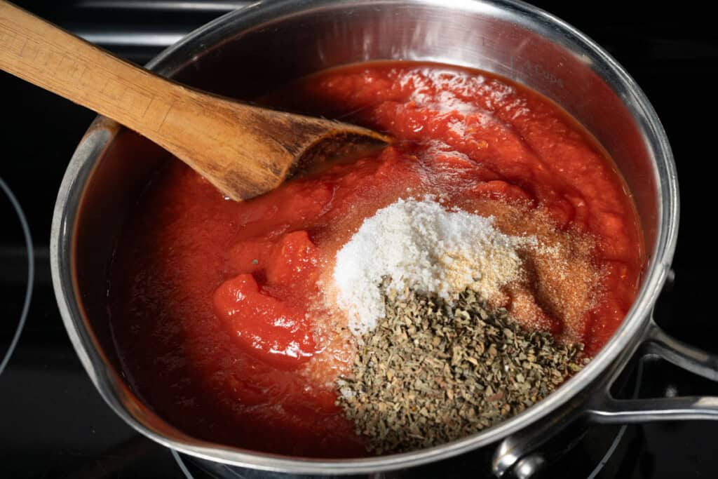 Adding crushed tomatoes and seasonings to a pot to make homemade pizza sauce.